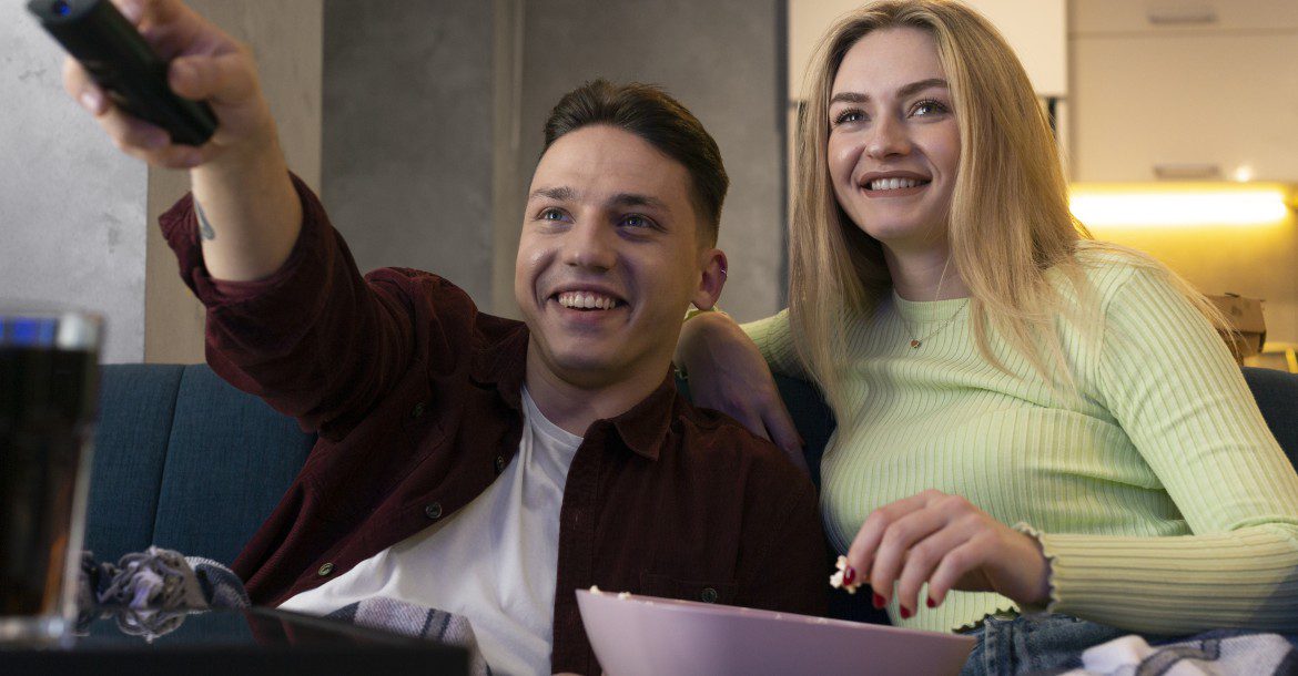 IPTV vs. Cable TV: Which One Wins in 2023? 12 A young man and woman sit on a couch with blankets, watching TV. The man holds a remote and points it at the screen while smiling. The woman holds a bowl of popcorn and laughs. They appear relaxed and happy, enjoying their favorite shows on Kemo TV.