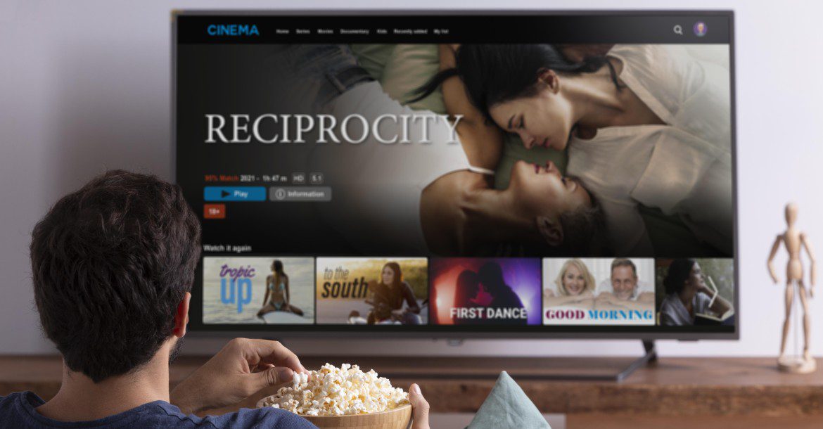 How to install IPTV? 12 A person holding a bowl of popcorn is watching Kemo IPTV on a large screen TV. The featured movie titled "Reciprocity" displays a couple lying down, with several other movie options listed below, including "Tropic Up," "In the South," "First Dance," and "Good Morning.