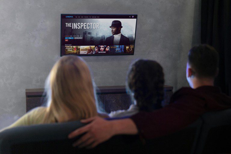 Three people are seated and watching a movie titled "The Inspector" on a wall-mounted TV in a dimly lit room. The man in the center has his arm around the person on his left. Various movie thumbnails, including options from KemoTV's IPTV channel list, are visible on the screen.