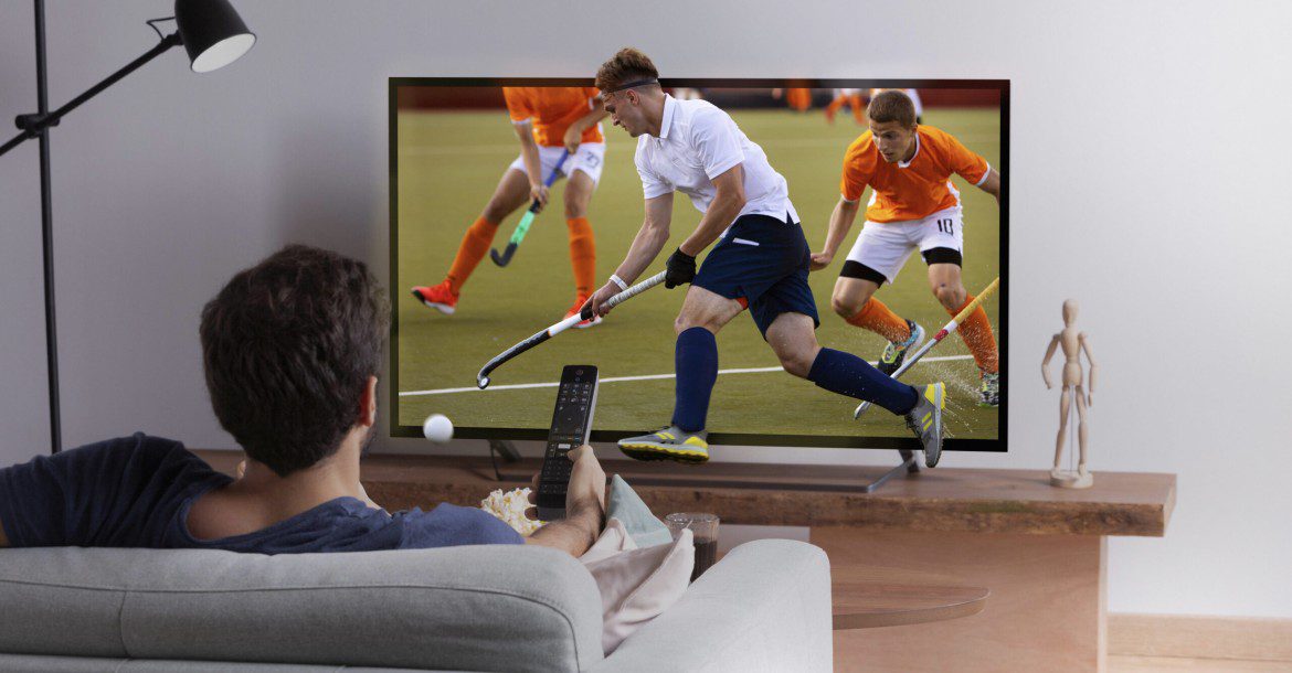 From HD to 8K: IPTV's Quest for Higher Resolutions in 2024 21 A person sits on a couch holding a remote control, watching a hockey game on Kemo IPTV displayed on a large flat-screen TV. The game features players in white and orange uniforms playing on a field. The living room has a modern design with a floor lamp and a small sculpture on a shelf.
