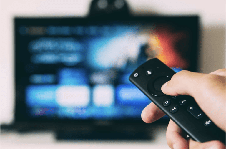 A person is holding a remote control in front of a blurred TV screen. The smart TV's interface displays app icons and a colorful background, indicating the kemoiptv home screen. The remote includes buttons for navigation, volume, and power.