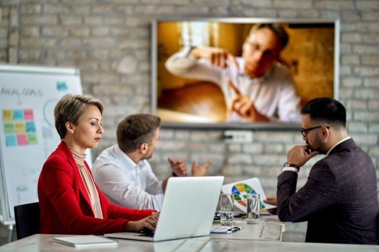 Three people are sitting at a conference table with laptops and documents, engaging in a video call with a person displayed on a large screen. The brick wall in the background features a whiteboard with colorful sticky notes and writing. The team uses Kemo IPTV to ensure seamless communication.