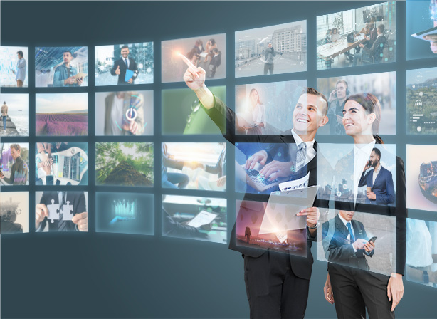 Two professionals, one pointing at a screen, stand in front of a large display featuring numerous small digital images. The images show various business-related activities and landscapes, giving the appearance of a high-tech work environment powered by KemoIPTV.