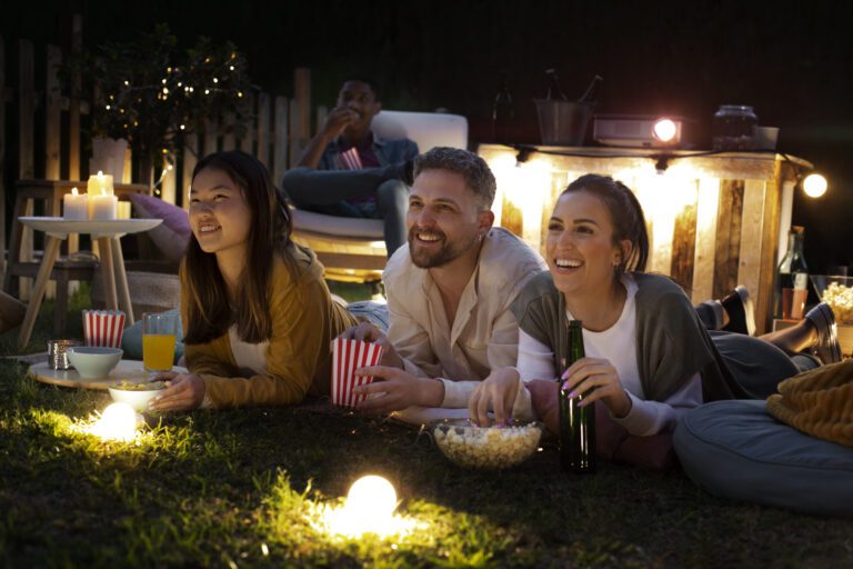 Three people are lying on a blanket on the grass at night, watching a projected film from Kemo TV. They are smiling, enjoying drinks, popcorn, and snacks. In the background, another person sits in a chair. Twinkling lights and a small table with candles create a cozy atmosphere.