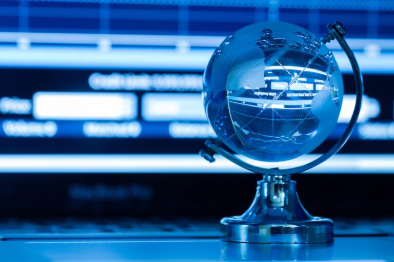 A small, metallic globe stands on a desk, illuminated by cool blue light. The globe reflects a blurred background that includes charts and graphs on a computer screen, hinting at global data or financial analysis—perhaps even KemoTV's latest insights from their IPTV channel list.