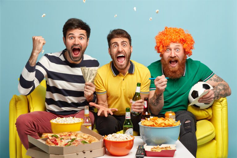 Three excited men watch a game on a yellow couch surrounded by snacks, pizza, and drinks. One man holds dollar bills, another holds multiple beer bottles, and the third, wearing an orange wig, has a soccer ball. Popcorn is mid-air, adding to the festive atmosphere as they stream the match on Kemo IPTV.