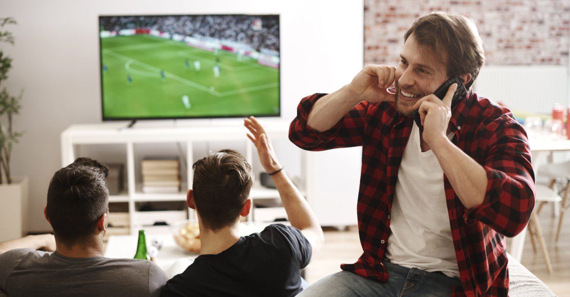 Exploring the Best IPTV Provider: Kemo IPTV 2 Three friends watch a soccer match on TV in a cozy living room. Two are seated on a couch, focused on the game from their KemoTV subscription, while the third stands with a smile, talking on two cellphones. The atmosphere looks lively and energetic.