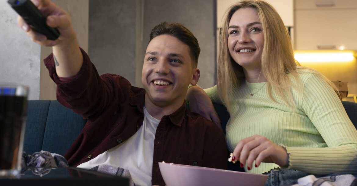 A young man and woman sit on a couch with blankets, watching TV. The man holds a remote and points it at the screen while smiling. The woman holds a bowl of popcorn and laughs. They appear relaxed and happy, enjoying their favorite shows on Kemo TV.