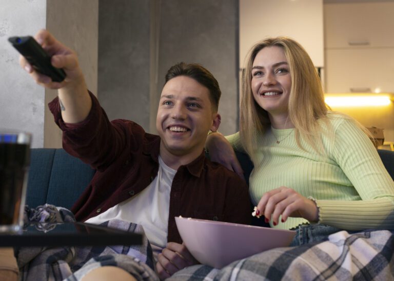 A young man and woman sit on a couch with blankets, watching TV. The man holds a remote and points it at the screen while smiling. The woman holds a bowl of popcorn and laughs. They appear relaxed and happy, enjoying their favorite shows on Kemo TV.