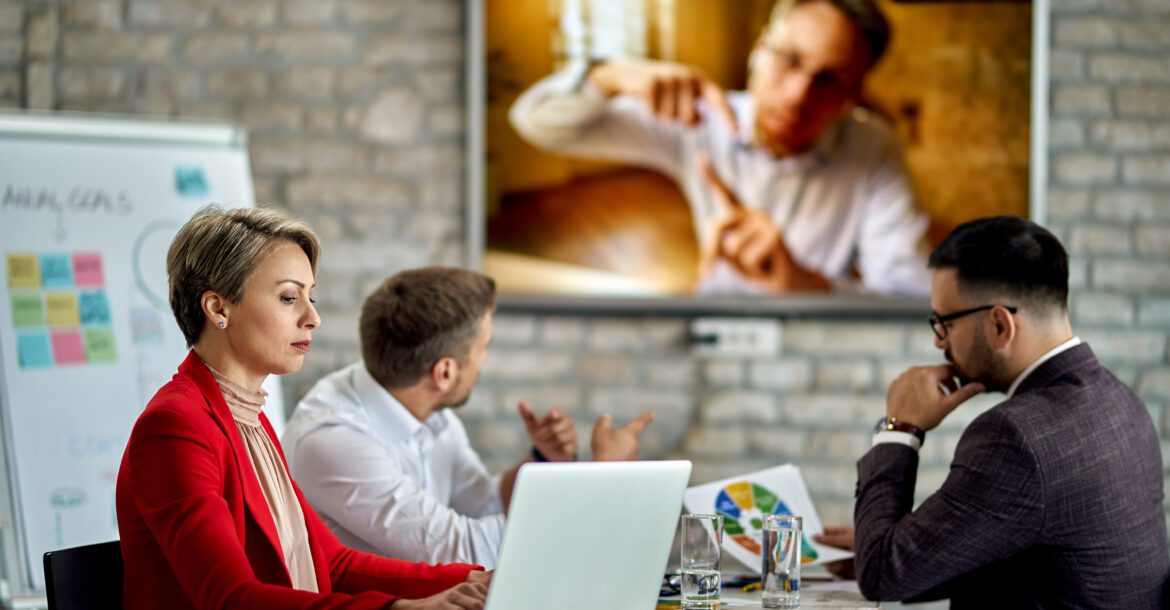 Three people are sitting at a conference table with laptops and documents, engaging in a video call with a person displayed on a large screen. The brick wall in the background features a whiteboard with colorful sticky notes and writing. The team uses Kemo IPTV to ensure seamless communication.