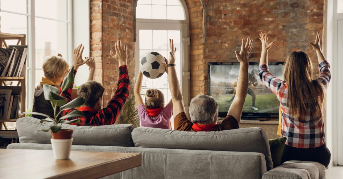 A group of people in plaid shirts, viewed from behind, sit on a gray couch in a rustic living room with exposed brick walls. They're watching a soccer match via Kemo IPTV and have their hands raised in celebration. A soccer ball is in mid-air.