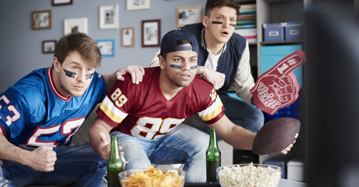 Three enthusiastic men watch a football game on TV in a living room. Two wear football jerseys, and one is holding a foam finger while the other grips a football. Snacks and drinks are on the coffee table. They seem intensely focused on the game, probably tuned into their favorite kemo iptv channel list.