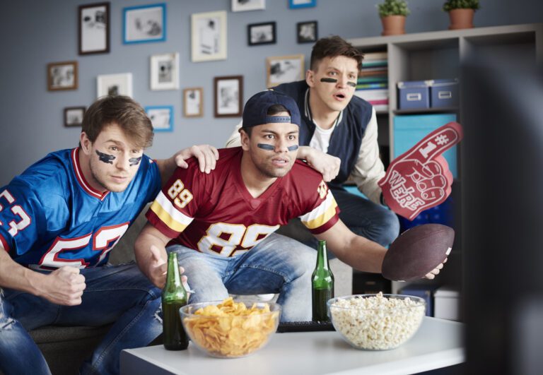 Three enthusiastic men watch a football game on TV in a living room. Two wear football jerseys, and one is holding a foam finger while the other grips a football. Snacks and drinks are on the coffee table. They seem intensely focused on the game, probably tuned into their favorite kemo iptv channel list.