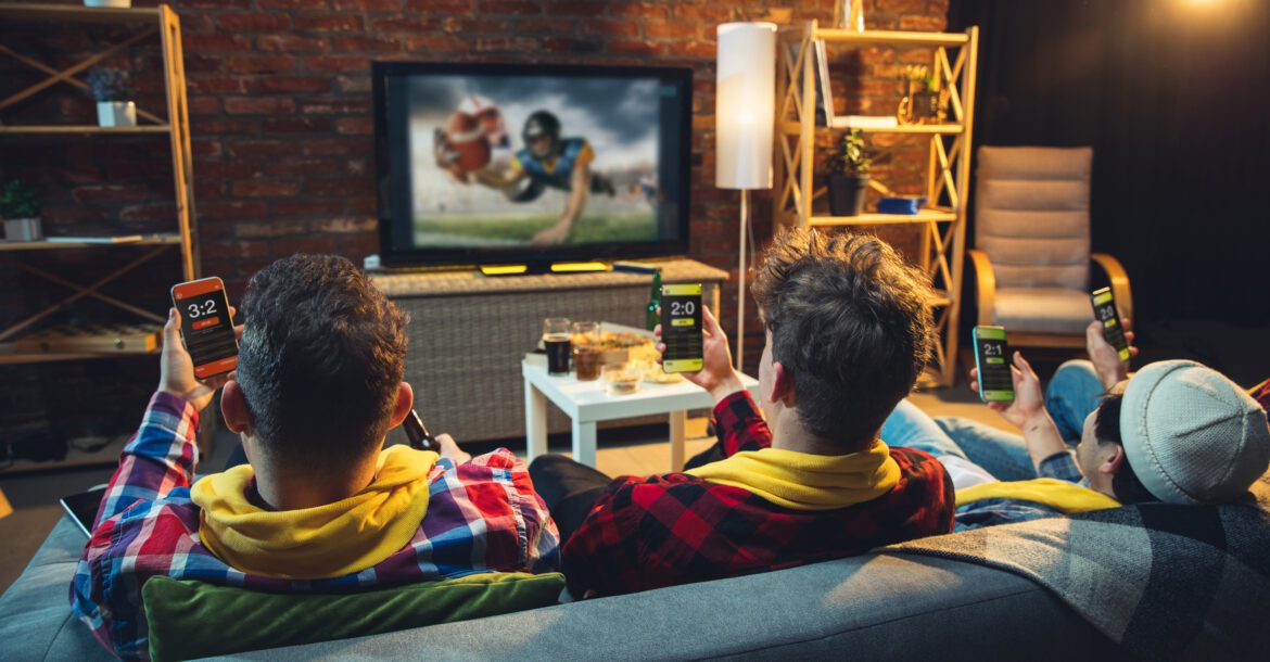 Three people sit on a couch watching a football game on KemoTV. They are holding smartphones displaying betting apps with live scores and stats. The room has a cozy setup with a brick wall, shelves, and a table with snacks and drinks.