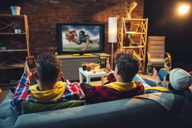 Three people sit on a couch watching a football game on KemoTV. They are holding smartphones displaying betting apps with live scores and stats. The room has a cozy setup with a brick wall, shelves, and a table with snacks and drinks.