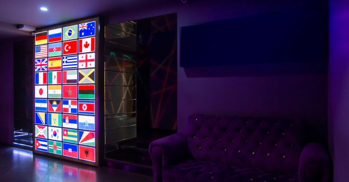 A dimly lit room with a wall displaying a collage of international flags illuminated by colorful lights, creating a kemoiptv vibe. To the right, there is a purple tufted sofa against a dark wall. The atmosphere appears modern and vibrant.