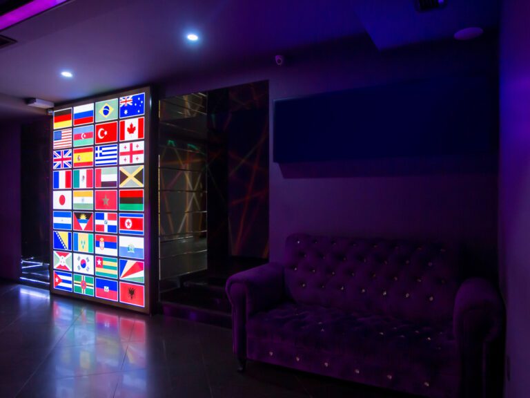 A dimly lit room with a wall displaying a collage of international flags illuminated by colorful lights, creating a kemoiptv vibe. To the right, there is a purple tufted sofa against a dark wall. The atmosphere appears modern and vibrant.