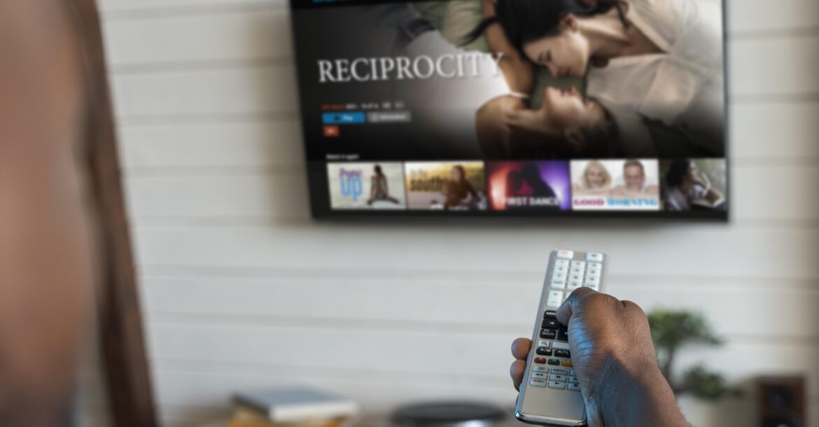 A person is pointing a remote control toward a flat-screen TV mounted on a white wall. The TV displays a streaming service interface with a highlighted movie titled "Reciprocity" featuring two people close together. Below the kemo tv, a media console is visible.