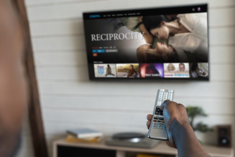 A person is pointing a remote control toward a flat-screen TV mounted on a white wall. The TV displays a streaming service interface with a highlighted movie titled "Reciprocity" featuring two people close together. Below the kemo tv, a media console is visible.