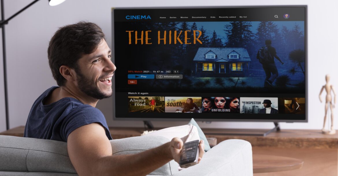 A smiling man sits on a couch holding a remote control, facing a TV screen displaying the kemoiptv interface. The highlighted movie is titled "The Hiker," featuring an image of a cabin in the woods and a person hiking. Other movie options from the kemo iptv channel list are visible below.