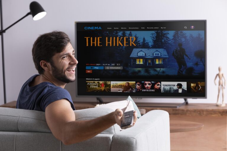 A smiling man sits on a couch holding a remote control, facing a TV screen displaying the kemoiptv interface. The highlighted movie is titled "The Hiker," featuring an image of a cabin in the woods and a person hiking. Other movie options from the kemo iptv channel list are visible below.