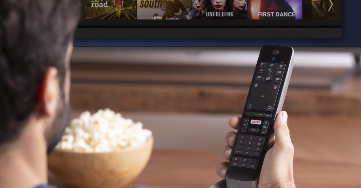 A person holding a remote control pointed at a Kemo TV screen. The TV displays a streaming service interface with various movie or TV show options. A bowl of popcorn is on the table in front of the person, who appears to be selecting something to watch on Kemotv.