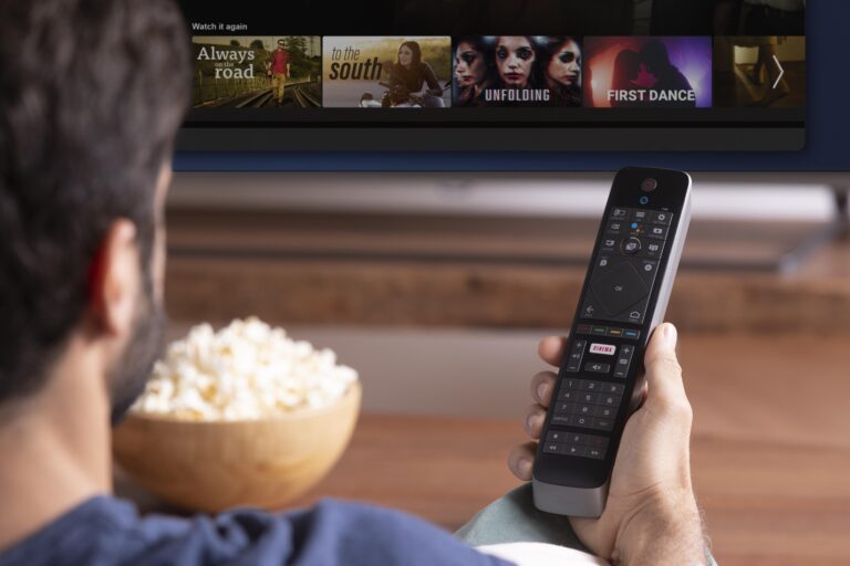 A person holding a remote control pointed at a Kemo TV screen. The TV displays a streaming service interface with various movie or TV show options. A bowl of popcorn is on the table in front of the person, who appears to be selecting something to watch on Kemotv.