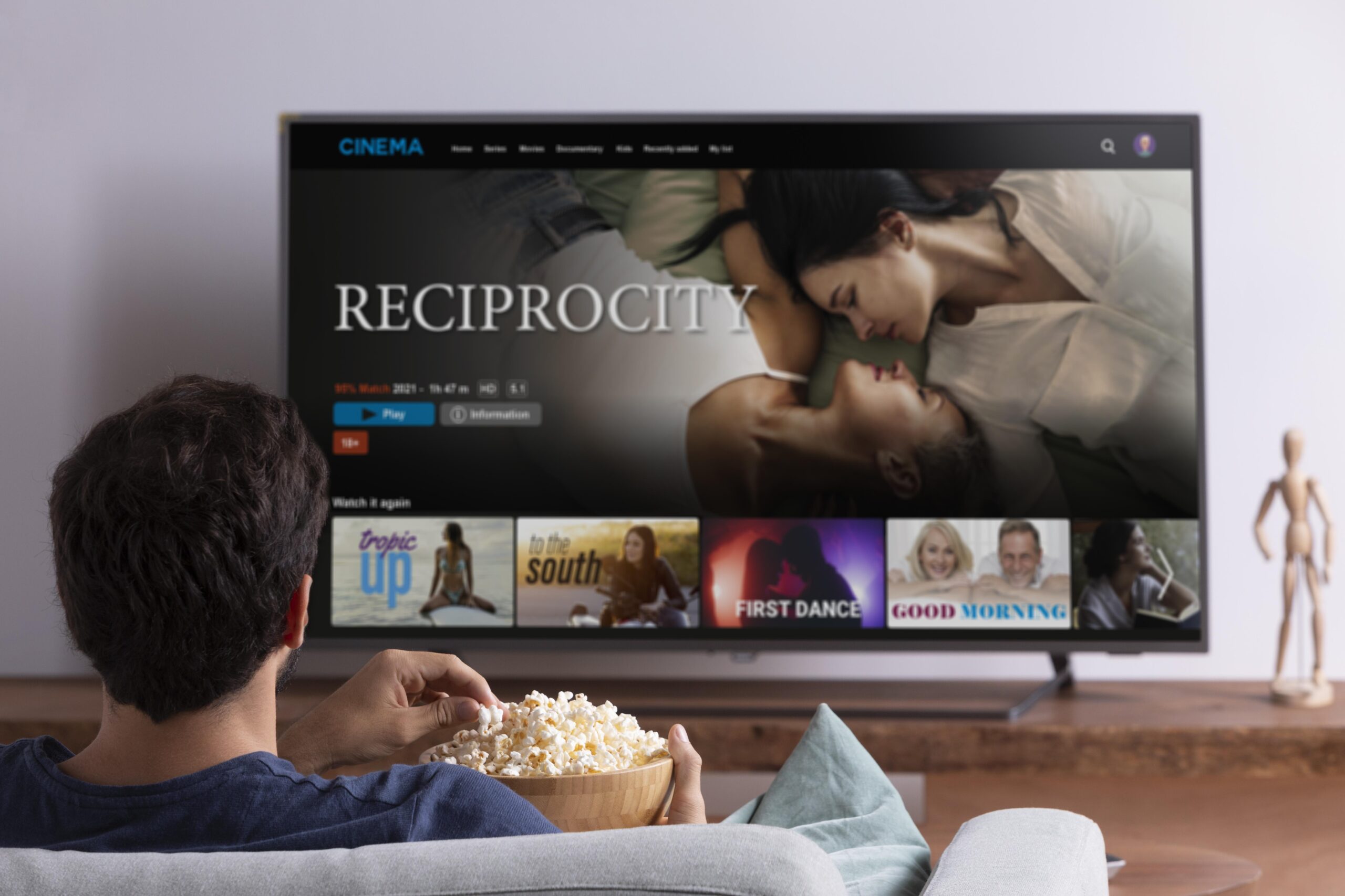 A person holding a bowl of popcorn is watching Kemo IPTV on a large screen TV. The featured movie titled "Reciprocity" displays a couple lying down, with several other movie options listed below, including "Tropic Up," "In the South," "First Dance," and "Good Morning.