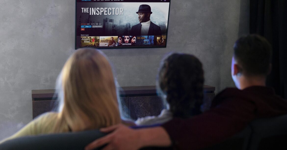 Three people are seated and watching a movie titled "The Inspector" on a wall-mounted TV in a dimly lit room. The man in the center has his arm around the person on his left. Various movie thumbnails, including options from KemoTV's IPTV channel list, are visible on the screen.