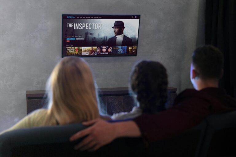 Three people are seated and watching a movie titled "The Inspector" on a wall-mounted TV in a dimly lit room. The man in the center has his arm around the person on his left. Various movie thumbnails, including options from KemoTV's IPTV channel list, are visible on the screen.