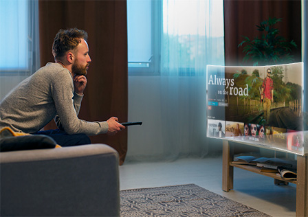 A man sits on a sofa in a modern living room, holding a remote control and watching a large wall-mounted TV. The screen displays the title "Always on the Road" with a scenic background image and additional smaller thumbnails beneath it, part of his Kemoiptv channel list.