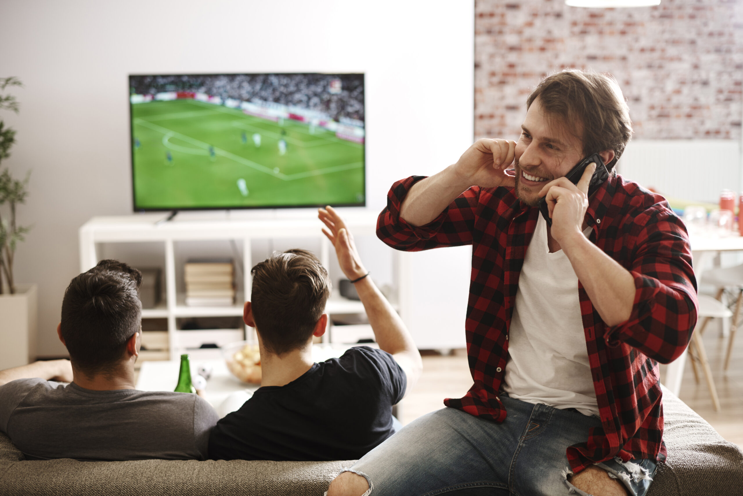 Three friends watch a soccer match on TV in a cozy living room. Two are seated on a couch, focused on the game from their KemoTV subscription, while the third stands with a smile, talking on two cellphones. The atmosphere looks lively and energetic.