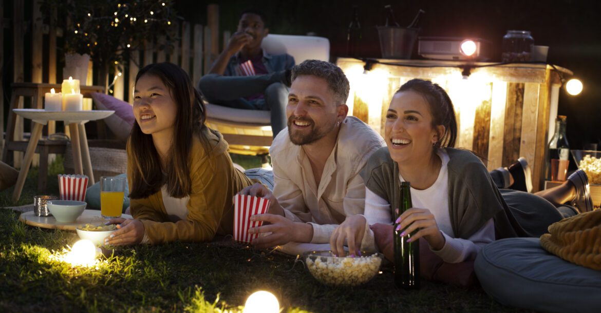 Three people are lying on a blanket on the grass at night, watching a projected film from Kemo TV. They are smiling, enjoying drinks, popcorn, and snacks. In the background, another person sits in a chair. Twinkling lights and a small table with candles create a cozy atmosphere.