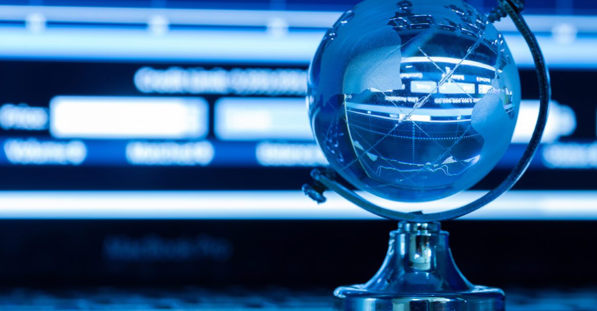 A small, metallic globe stands on a desk, illuminated by cool blue light. The globe reflects a blurred background that includes charts and graphs on a computer screen, hinting at global data or financial analysis—perhaps even KemoTV's latest insights from their IPTV channel list.