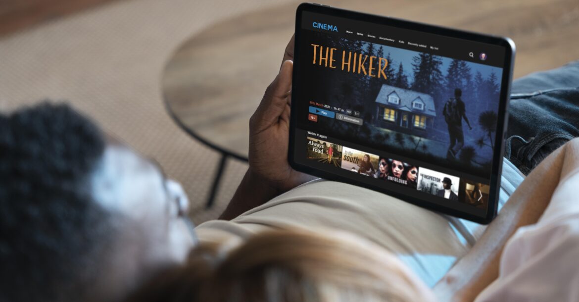 A man and woman are lying on a sofa, with the man holding a tablet. The tablet screen displays the KemoTV app, showing a film titled "The Hiker." The movie's cover depicts a haunted house and a figure walking in the woods.